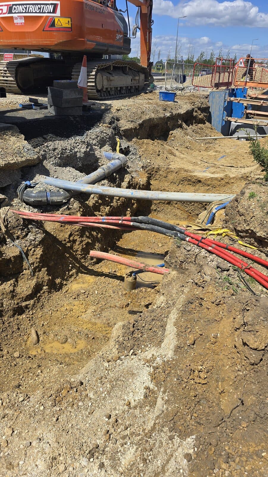 Excavator beside deep trench exposing grey water pipe and bundled red and black electrical cables at a construction site
