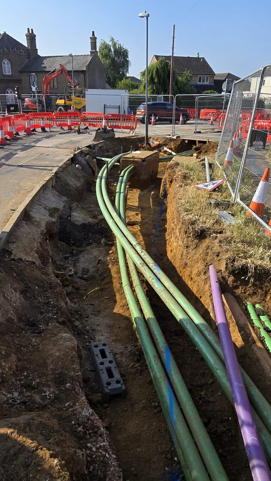 Long trench with green and purple ducting pipes exposed near roadside houses and orange traffic cones