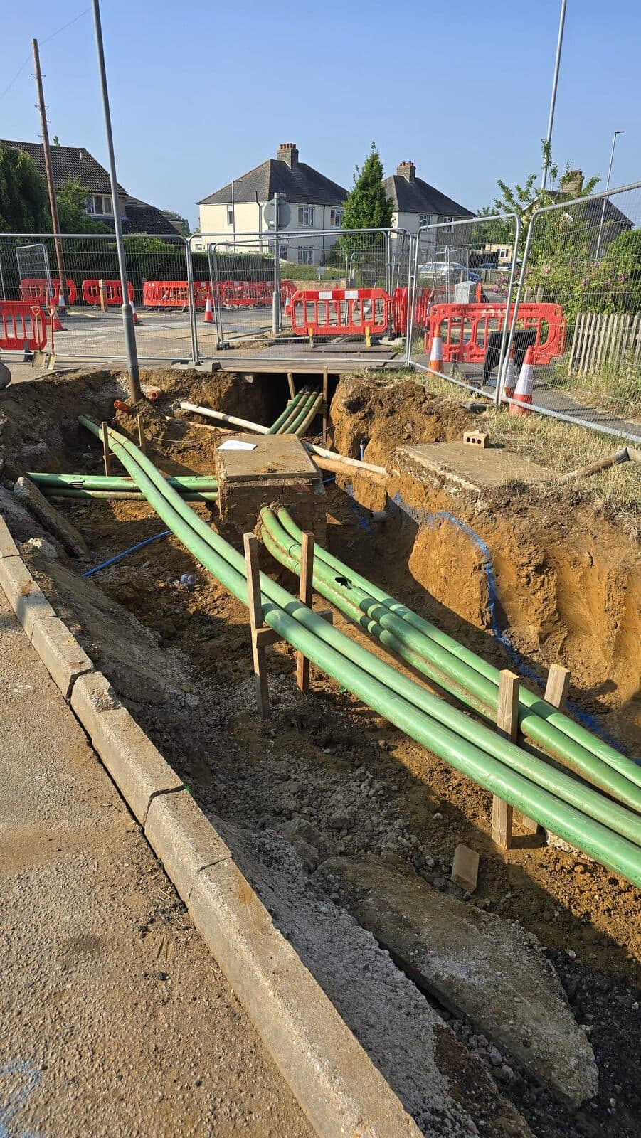 Trench with green ducting pipes supported by wooden stakes near residential street with red fencing barriers
