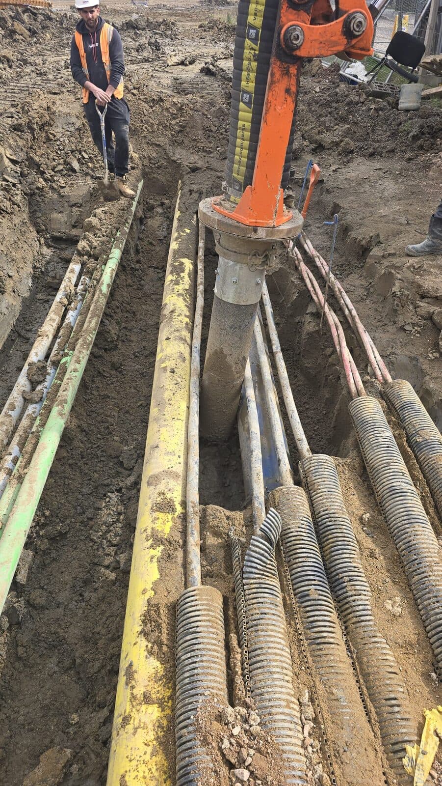 Worker standing beside deep trench with exposed yellow gas pipe and bundled orange ducting, vacuum excavation equipment visible