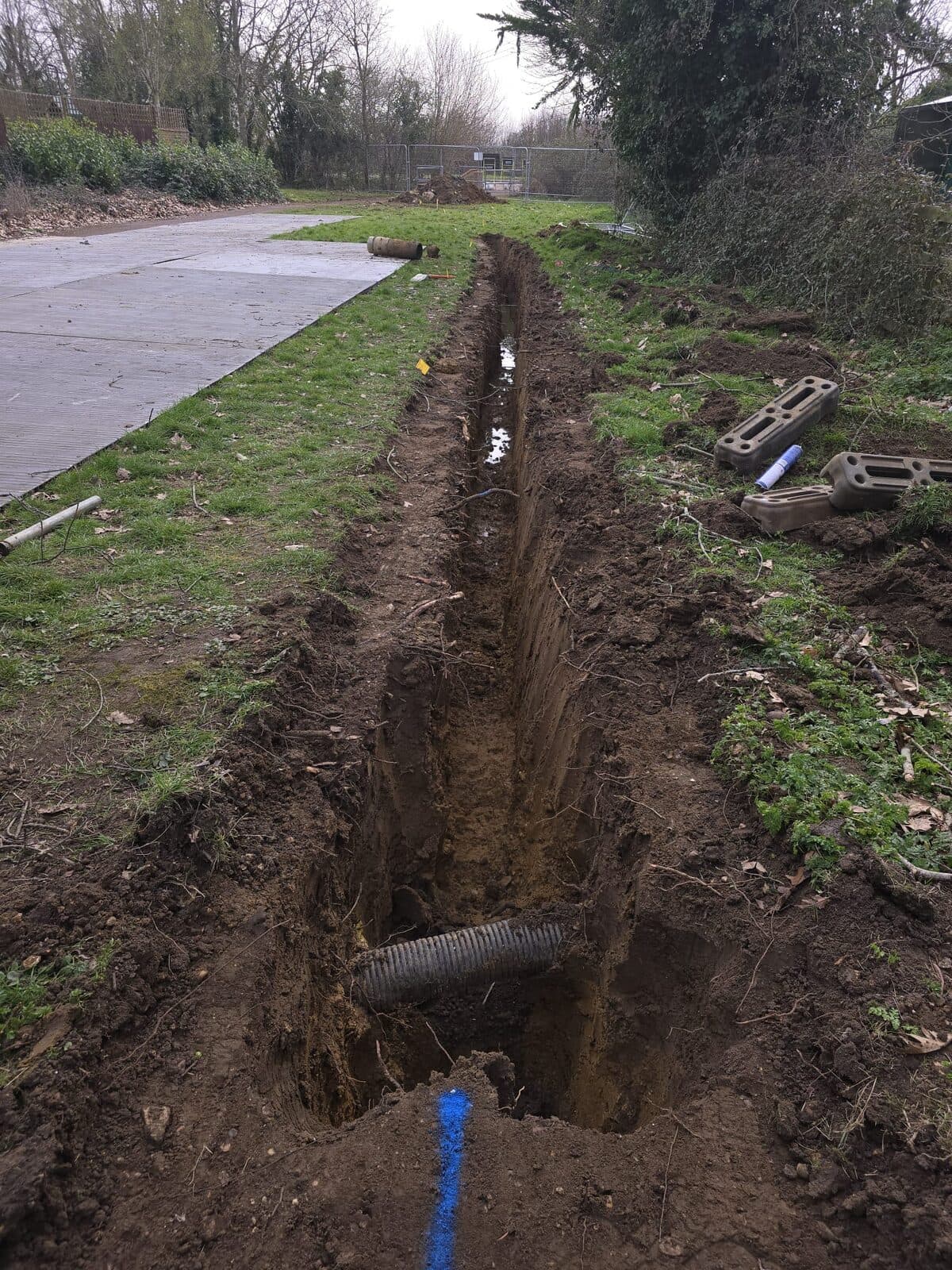 Long empty excavated trench with exposed black ducting pipe running through grassy area near parking lot