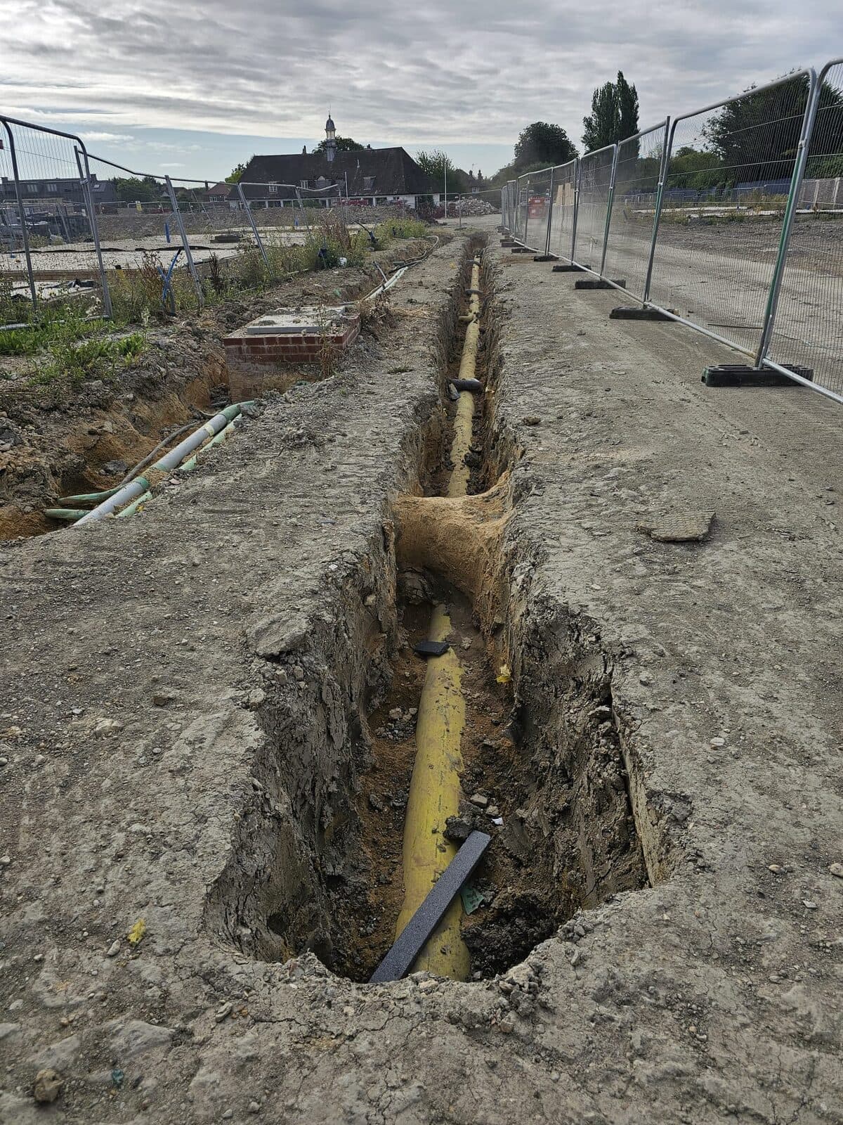 Deep straight trench with exposed yellow gas pipe running along fenced construction site under cloudy sky