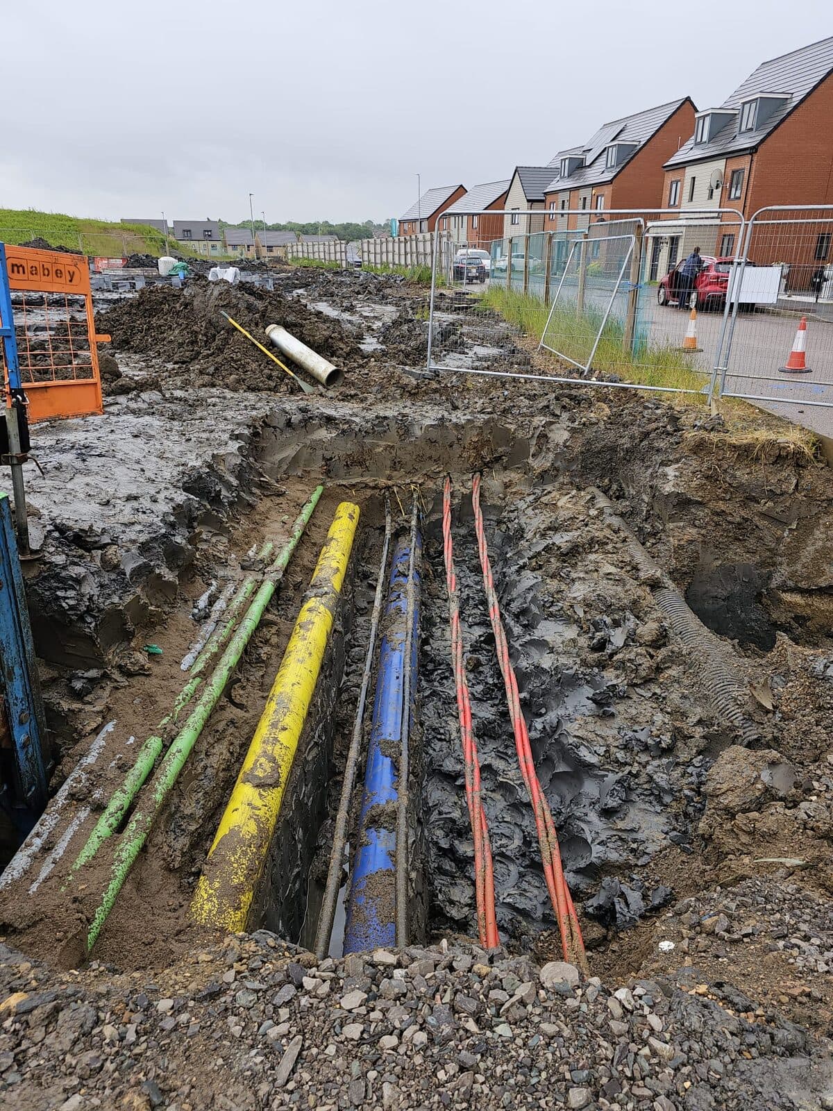 Trench exposing yellow, blue, and orange cables in muddy pit near residential housing development